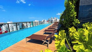 Una piscina infinita en la azotea con sillones de descanso, vistas panorámicas del horizonte urbano y exuberante vegetación.