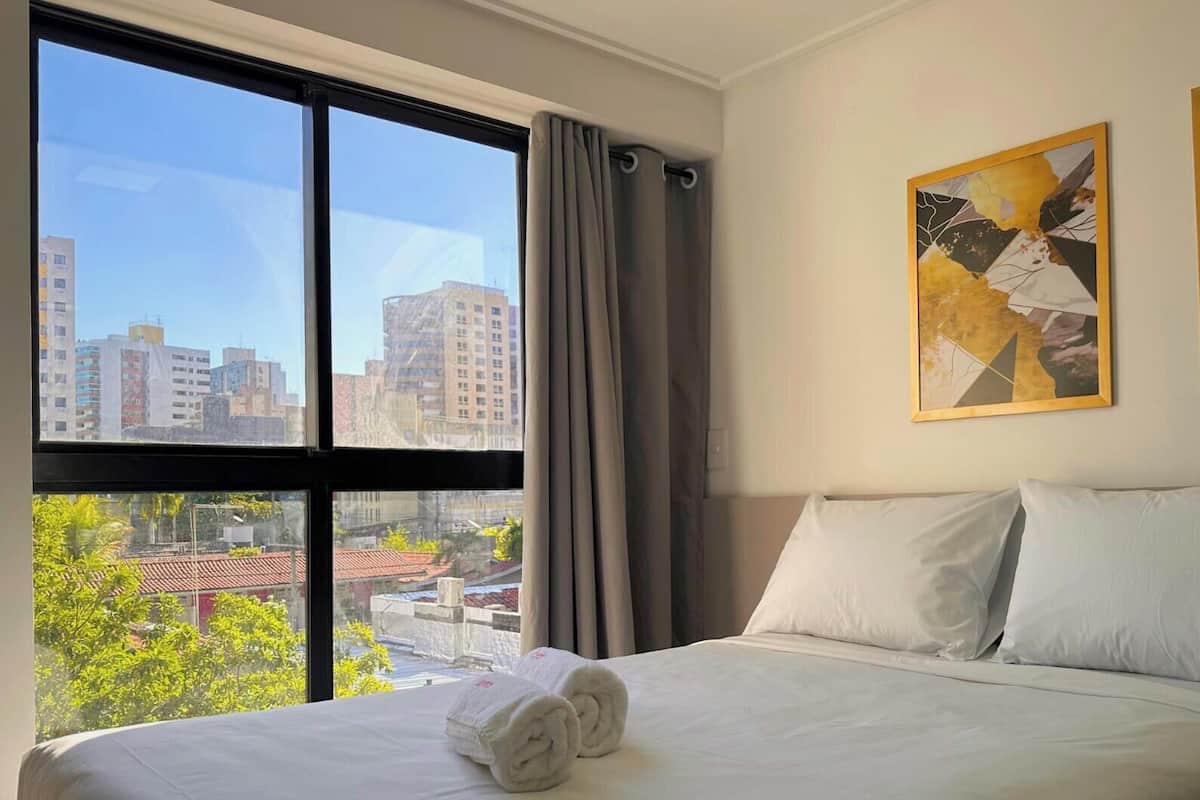 Habitación de hotel con un amplio ventanal, una cama con ropa blanca y dos cuadros abstractos en la pared.