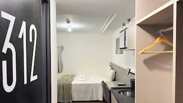 A compact room with a bed, a small table, a hanging clothes rack, and a sink.