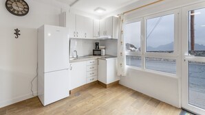 Cozinha com armários brancos, geladeira, microondas e cafeteira. Há um relógio na parede e uma janela com vista para o exterior.
