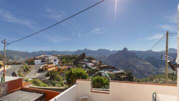 Un balcón con vistas a un pueblo en la ladera, cielo despejado y montañas a lo lejos.