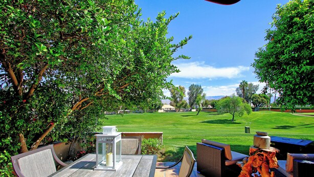 An outdoor patio with a table and chairs, a decorative tree stump, and a view of a well-maintained lawn and distant buildings. - 2 Bedroom at Woodhaven Country Club in Palm Desert. (Palm Desert)
