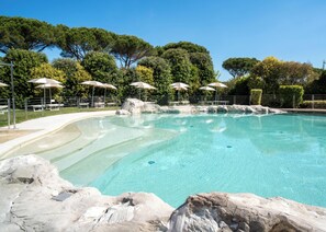 A clear swimming pool with rock formations, surrounded by trees and umbrellas. - Hotiday Lodge Viareggio (Viareggio)