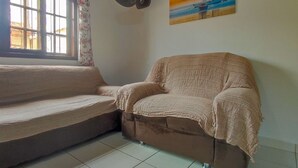 A living room with a sofa and a chair, a window with curtains, a wall-mounted fan, and a painting of a sunset over the sea. - Cozy house in Balneário Jussara, Mongaguá. Just 600m from the beach. Pets allowed. (Mongaguá)