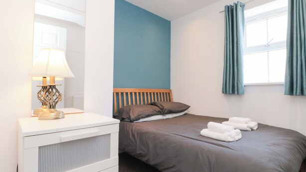 A bedroom with a bed, a bedside table with a lamp, a window with curtains, and a radiator. - Farriers Cottage (Bridlington)
