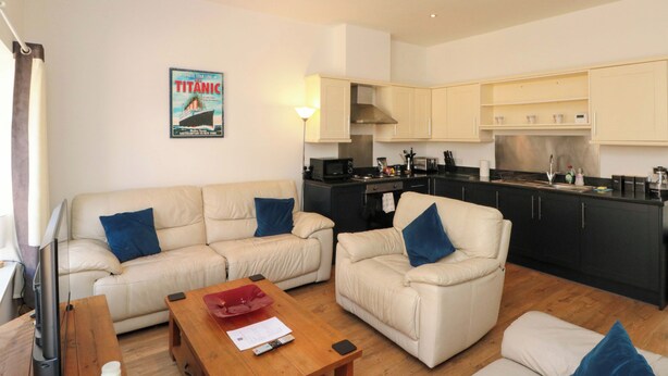 A living room with a sofa and armchair, a wooden coffee table, a kitchen with cabinets and appliances, and a poster on the wall. - Chestnut Cottage (Bridlington)