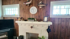 Ein Wohnzimmer mit Kamin, Holzvertäfelung, an Wänden befestigten Fischdekorationen, einer Uhr und einem Kaminvorsatz mit der Aufschrift 'HOME' und kleinen Pflanzen.