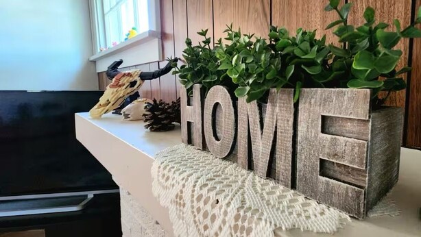Ein Holzblock mit dem Wort 'HOME' und einer Pflanze darauf, auf einer weißen Oberfläche mit einem gewebten Untersetzer.