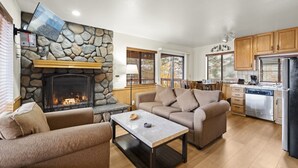 Un salon chaleureux avec une cheminée en pierre, un canapé, une table basse et un coin cuisine doté de meubles en bois et d’un réfrigérateur.