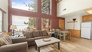 A spacious living area with a large window overlooking a body of water, a cozy seating arrangement, and a kitchen area with wooden cabinets. - 2BR Tahoe Snowfall, Lake Mist & Crackling Fires (Tahoe Vista)