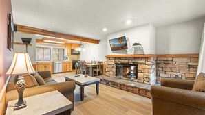 A spacious living area with a stone fireplace, wooden furniture, and a kitchen in the background. - 3BR Basecamp for Tahoe Days Done Right (Tahoe Vista)