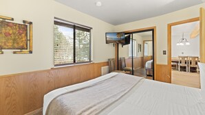 A bedroom with a bed, a window with blinds, a painting on the wall, and a view of a dining area. - 3BR Basecamp for Tahoe Days Done Right (Tahoe Vista)