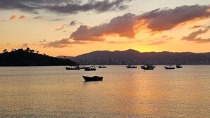 Un plan d’eau calme où flottent plusieurs bateaux, avec, au loin, la silhouette d’une ville, et un coucher de soleil aux couleurs flamboyantes dans un ciel nuageux.