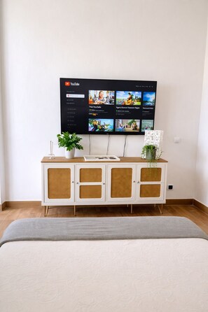 Ein Schlafzimmer mit einem Bett, einem weißen Schminktisch mit Pflanze und einem an der Wand montierten Fernseher, der YouTube-Kanäle anzeigt.