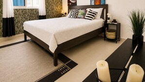 Un dormitorio con cama, mesita de noche, escritorio con velas, planta en maceta y ventana con cortinas.