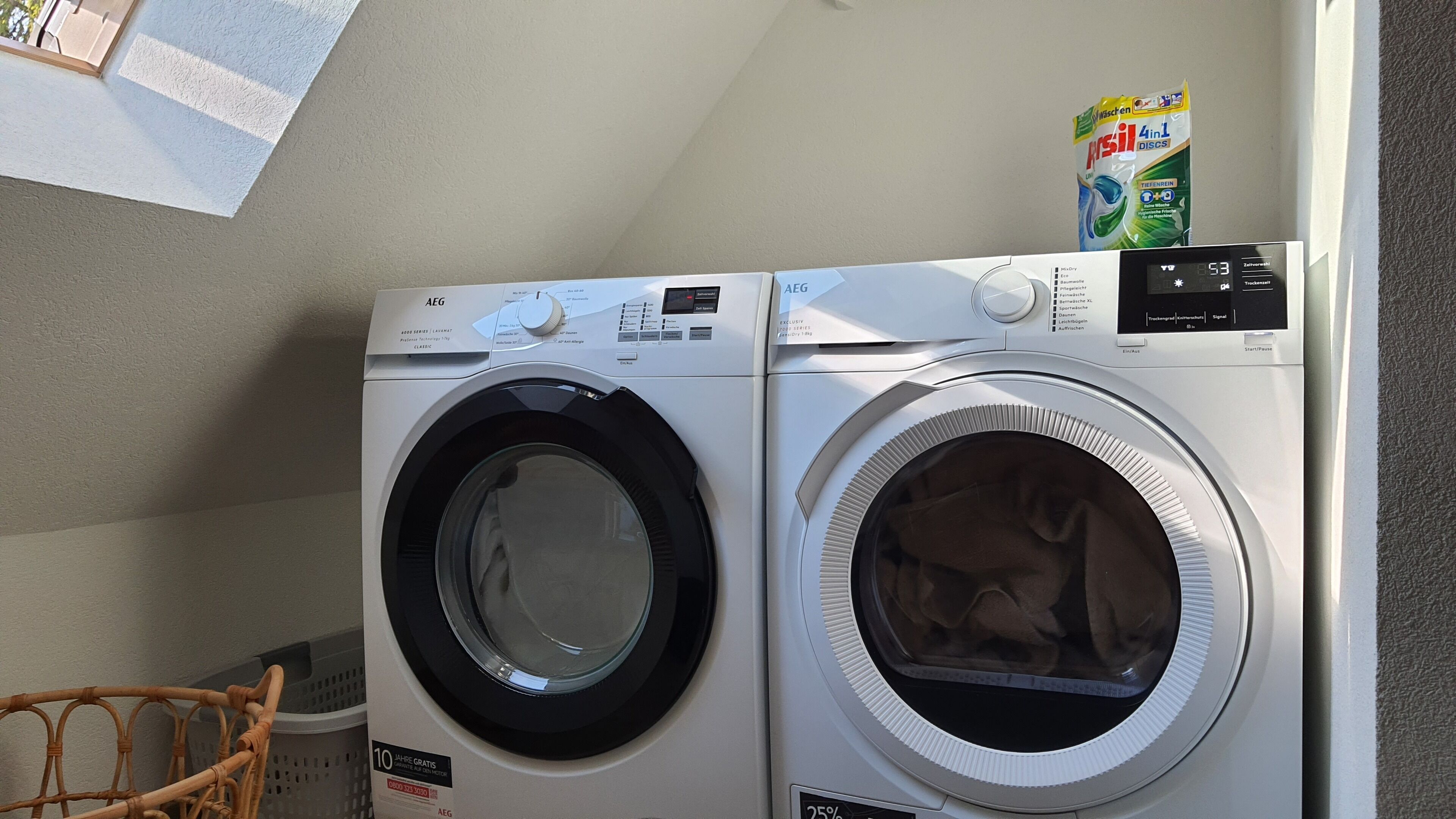 A front-loading washing machine and dryer in a laundry room with a basket and detergent visible.