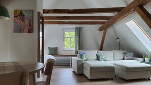 A cozy loft-style room with exposed wooden beams, a sofa set, a dining table, and a window with curtains. - Fewo am Kirchbrunnen (DG) - Quiet time out for 2 (Urspringen vor der Rhön)