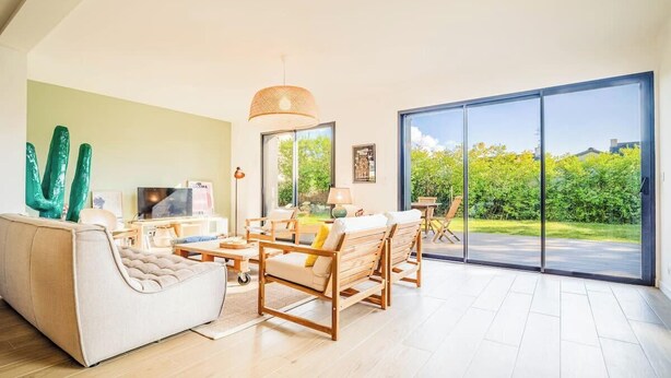 A spacious living room with a sofa, wooden chairs, a TV, and a large sliding glass door leading to a garden. - Belle maison moderne proche plages & bourg (Carnac)