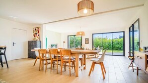A spacious dining area with a large wooden table and chairs, a hanging light fixture, and a view of greenery through large windows. - Belle maison moderne proche plages & bourg (Carnac)