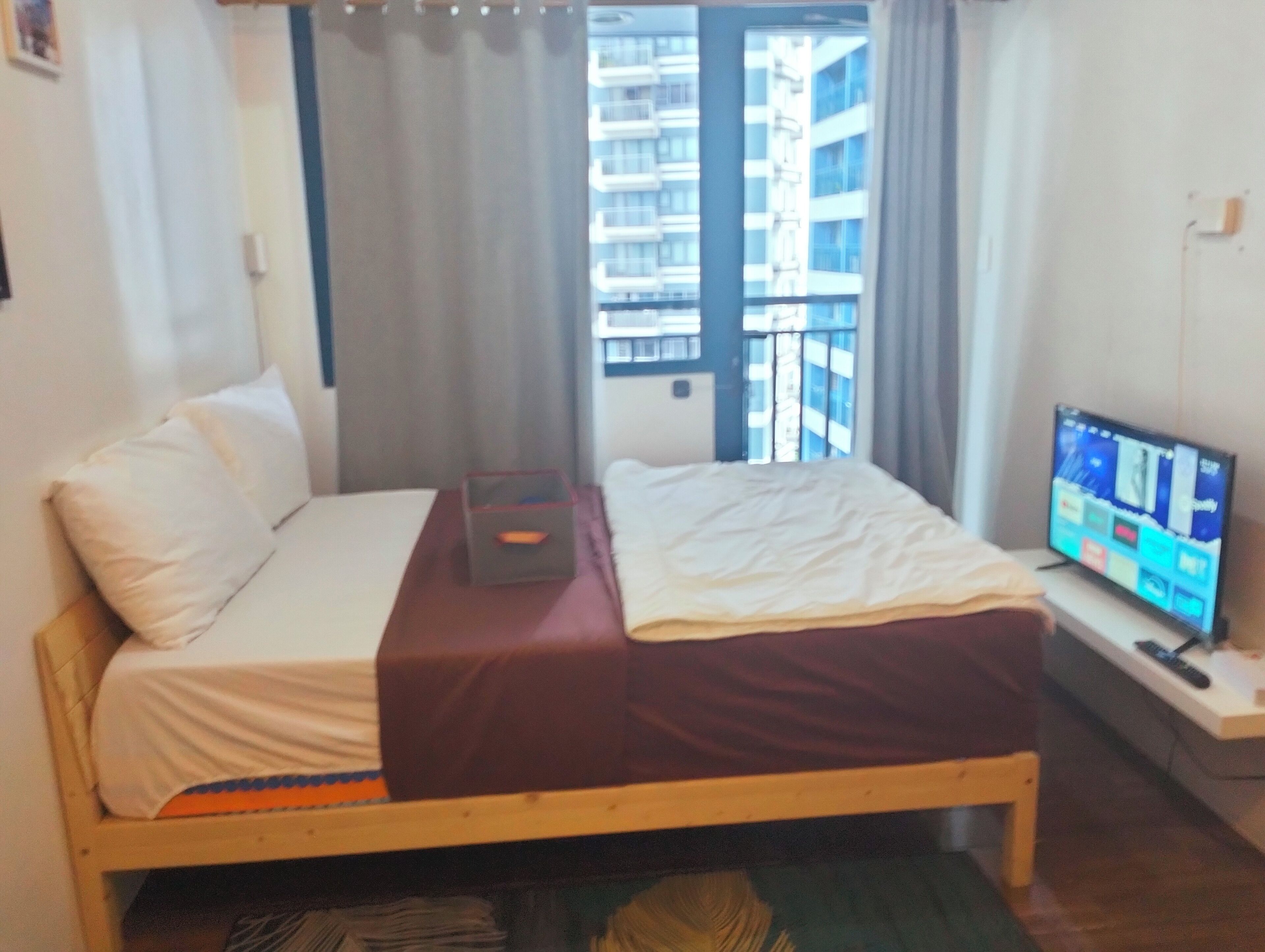 A bedroom with a bed, a TV, a window with curtains, and a view of buildings outside.