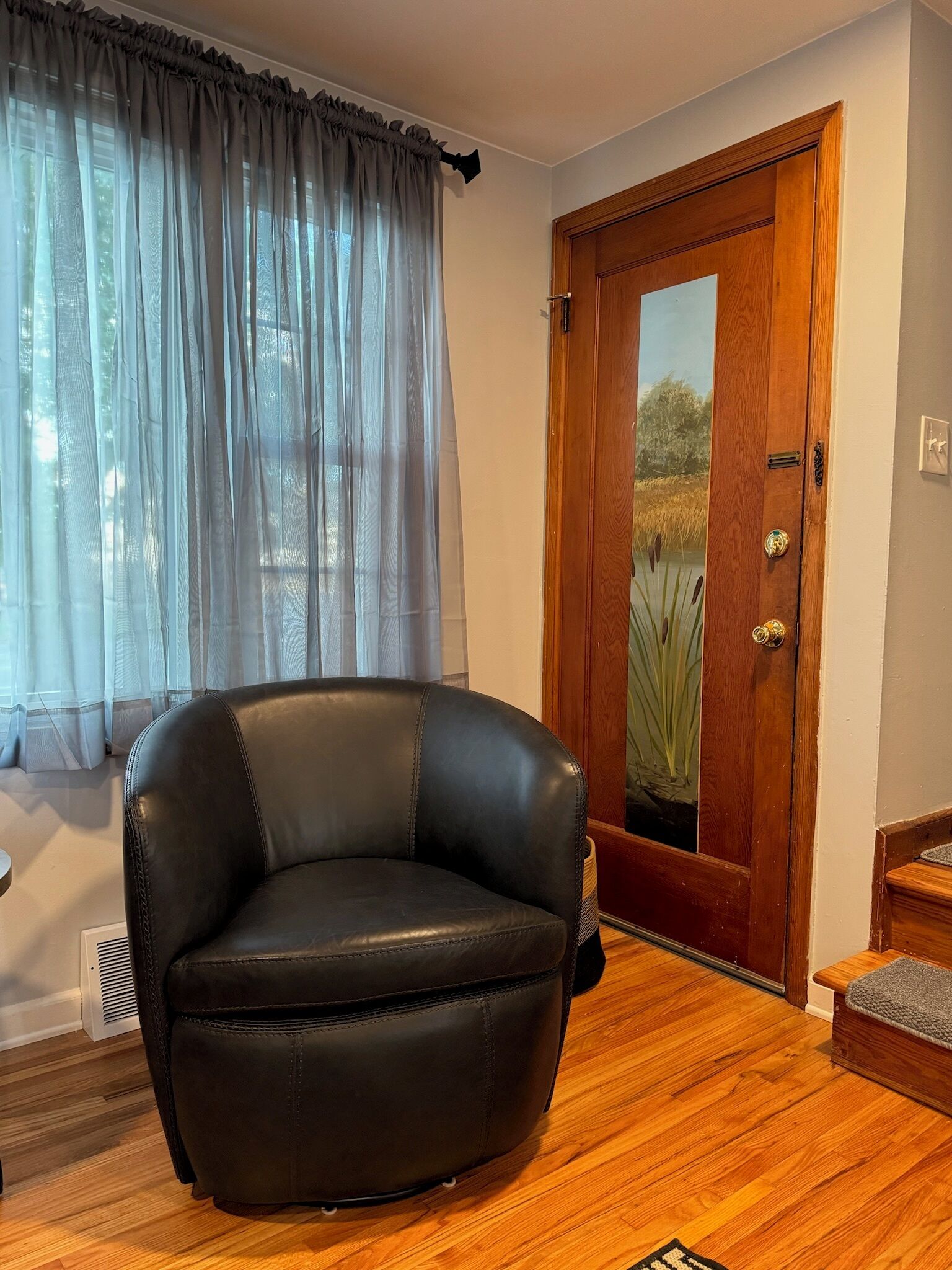 Une pièce avec un fauteuil en cuir noir, un plancher en bois, une porte en bois dotée d’un panneau de verre et des rideaux légers.