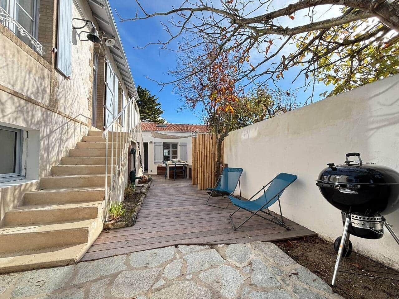 Een achtertuin met een pad van stenen dat leidt naar een houten terras, een barbecue en blauwe klapstoelen.