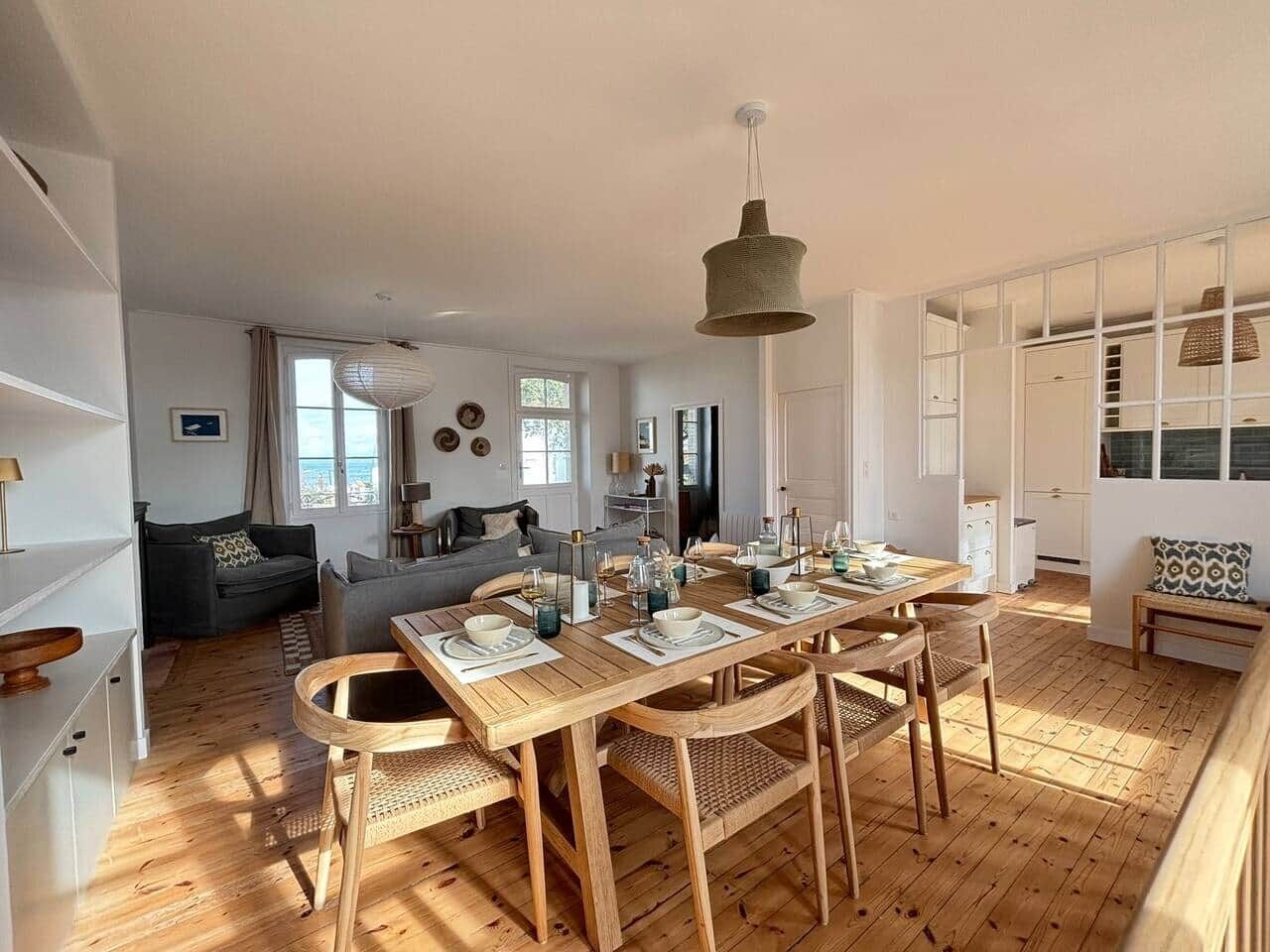 Un vaste espace de vie comprend une table à manger en bois, préparée pour accueillir quatre personnes, entourée de chaises en osier. Une grande fenêtre laisse entrer la lumière naturelle, et un canapé est placé contre le mur.