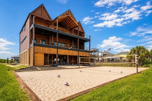 A two-story wooden building with a balcony, a sandy volleyball court, and a grassy area. - Beach Front | Hot Tub | Volleyball | Modern Construction (Crystal Beach)