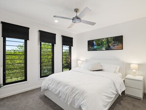 Une chambre à coucher avec un grand lit, deux tables de chevet, un ventilateur de plafond et trois fenêtres équipées de stores noirs.