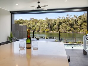Un espace de repas moderne avec une table, deux verres et une bouteille de vin. Une grande fenêtre offre une vue sur un lac et des arbres.