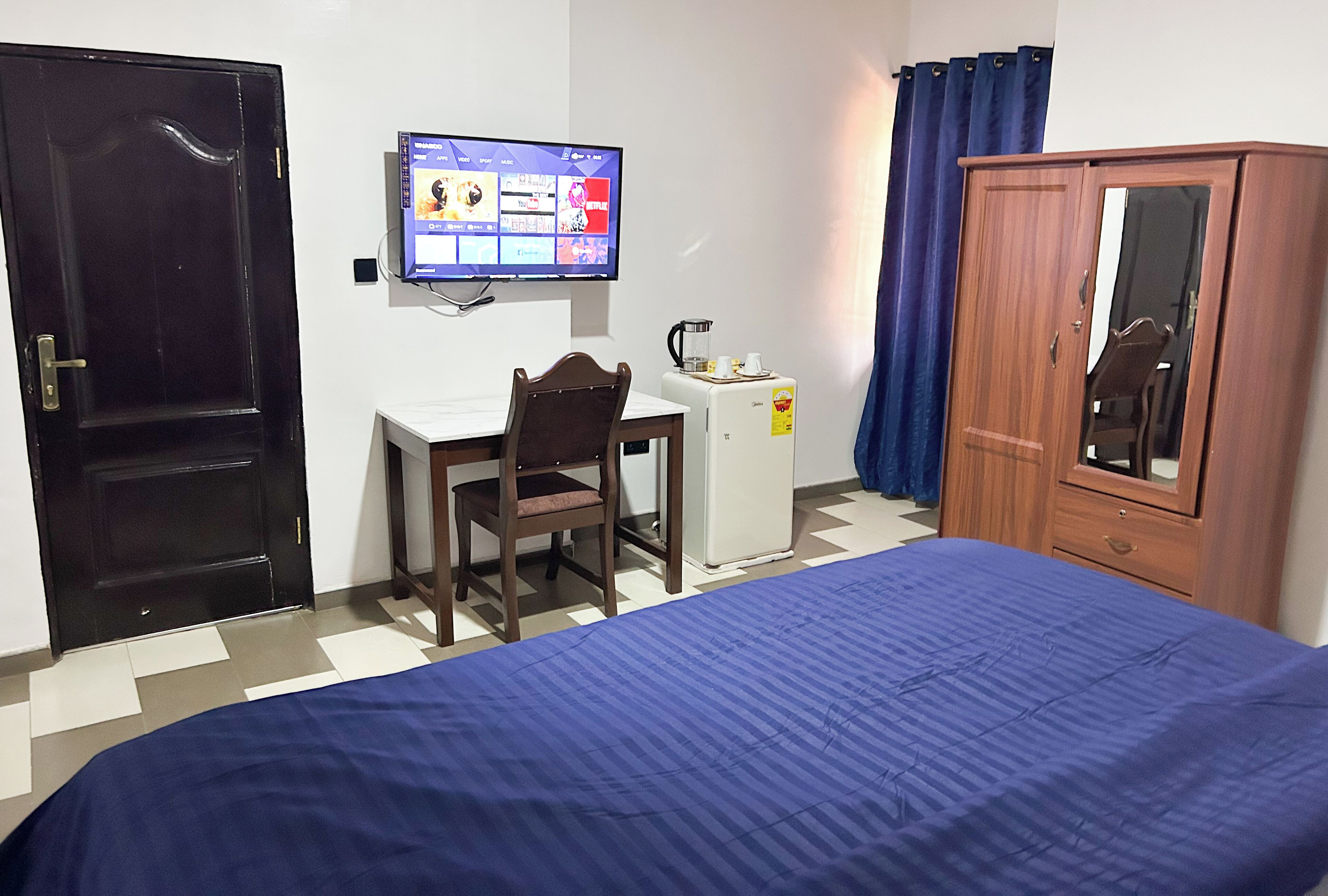 A bedroom with a bed, a desk with a chair, a television, a wardrobe, and a small refrigerator.