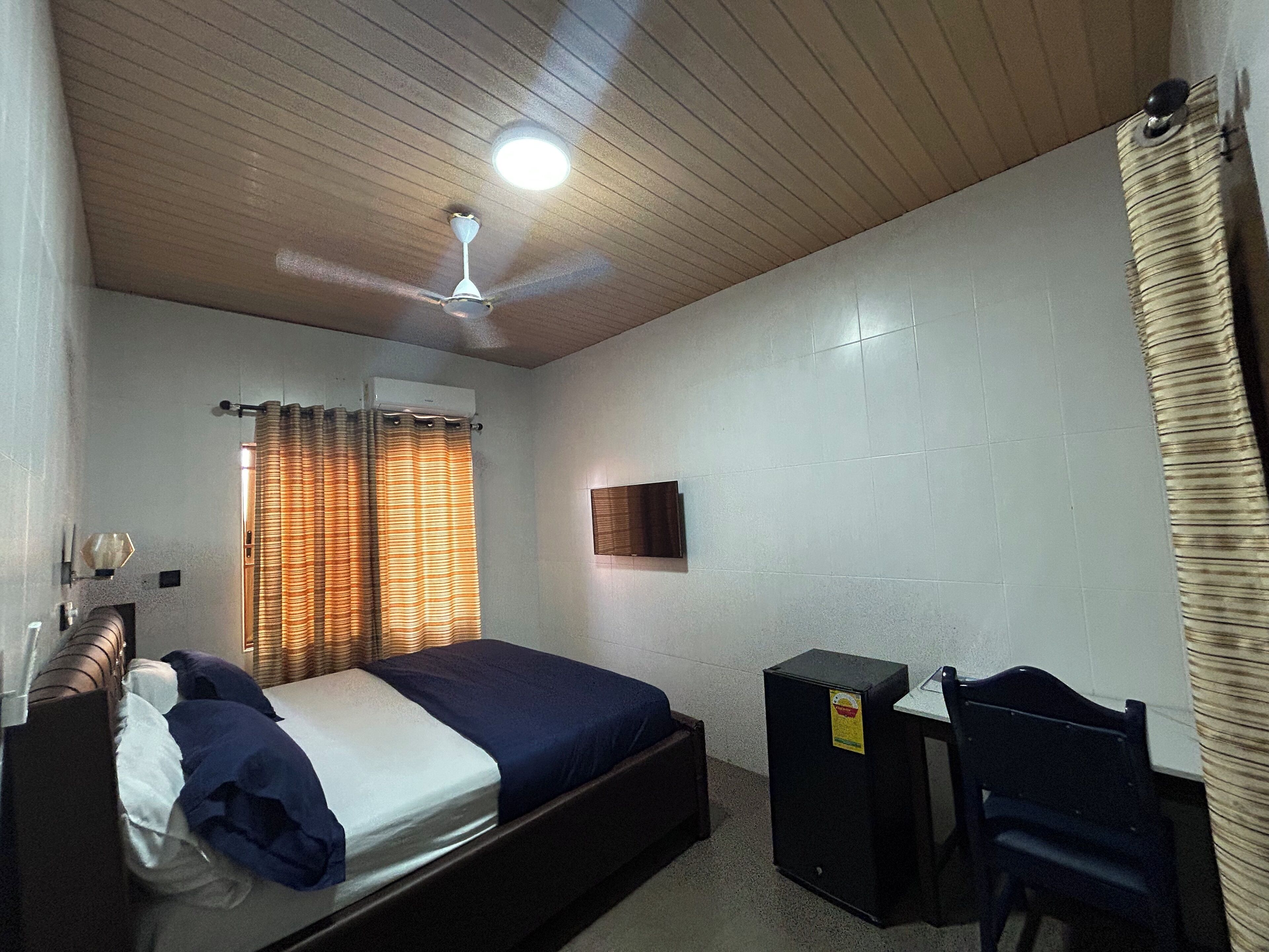 Une chambre à coucher comprenant un lit, un bureau, une chaise, un ventilateur de plafond et un téléviseur fixé au mur.