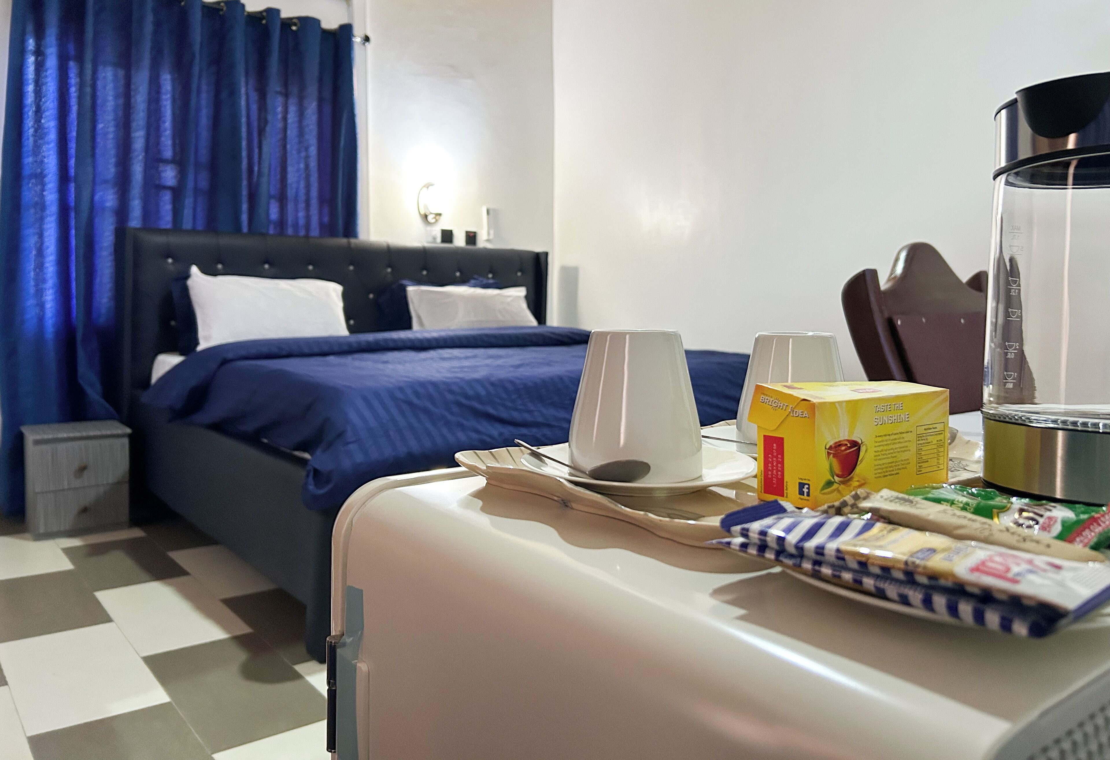 Habitación de hotel con cama, mesita de noche con cafetera, caja de pasteles y bandeja de desayuno.