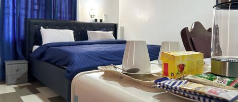 Habitación de hotel con cama, mesita de noche con cafetera, caja de pasteles y bandeja de desayuno.