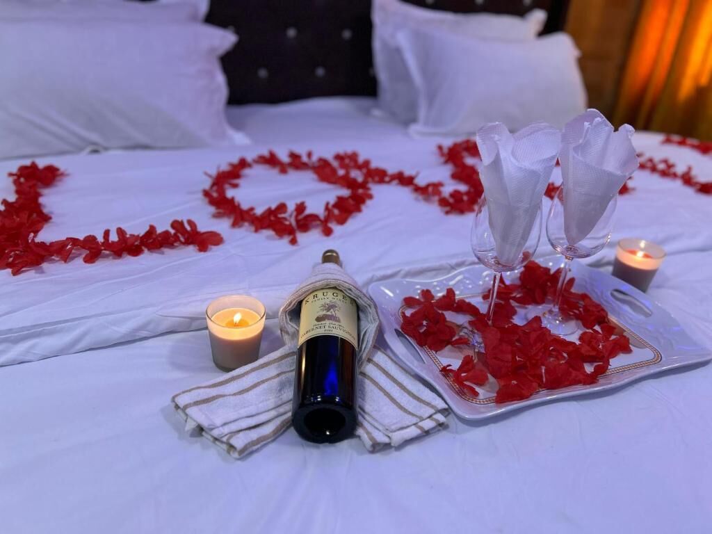 A bed with white linens, a bottle of wine, a candle, and red rose petals arranged in a heart shape.