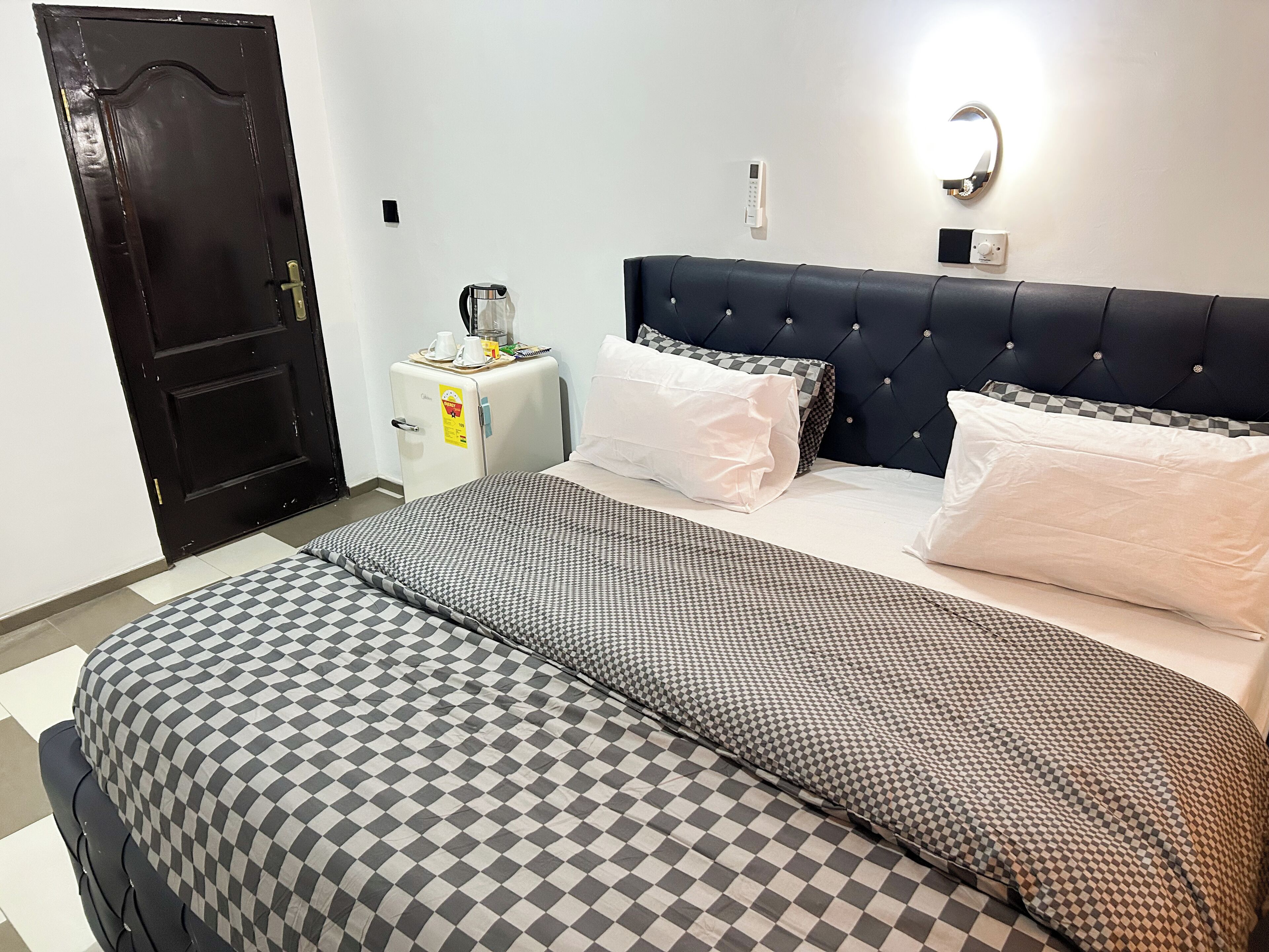 A hotel room with a checkered bedspread, a dark upholstered headboard, and a small bedside table with a kettle and a cup.
