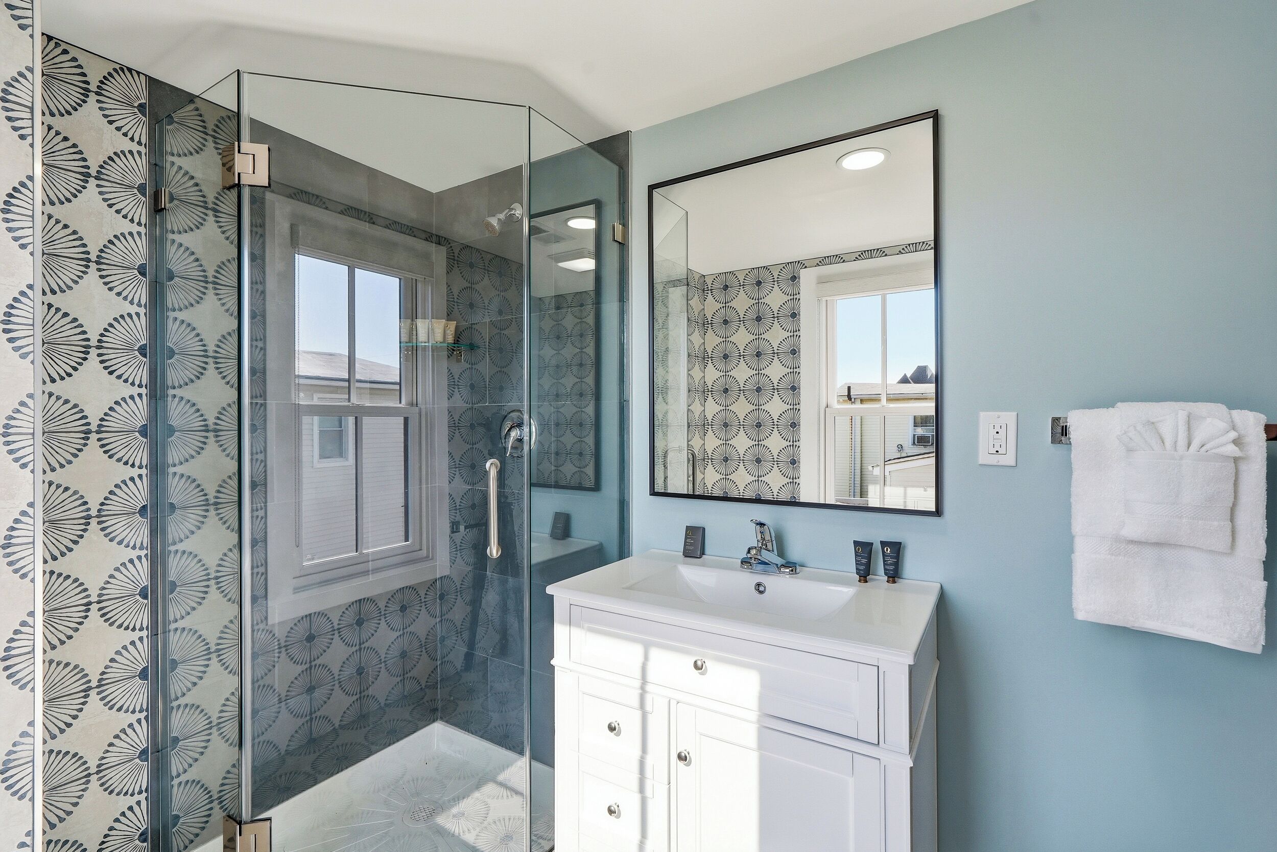 Une salle de bain dotée d’une douche à parois de verre, d’un meuble-lavabo blanc avec un miroir et de carreaux muraux à motifs.