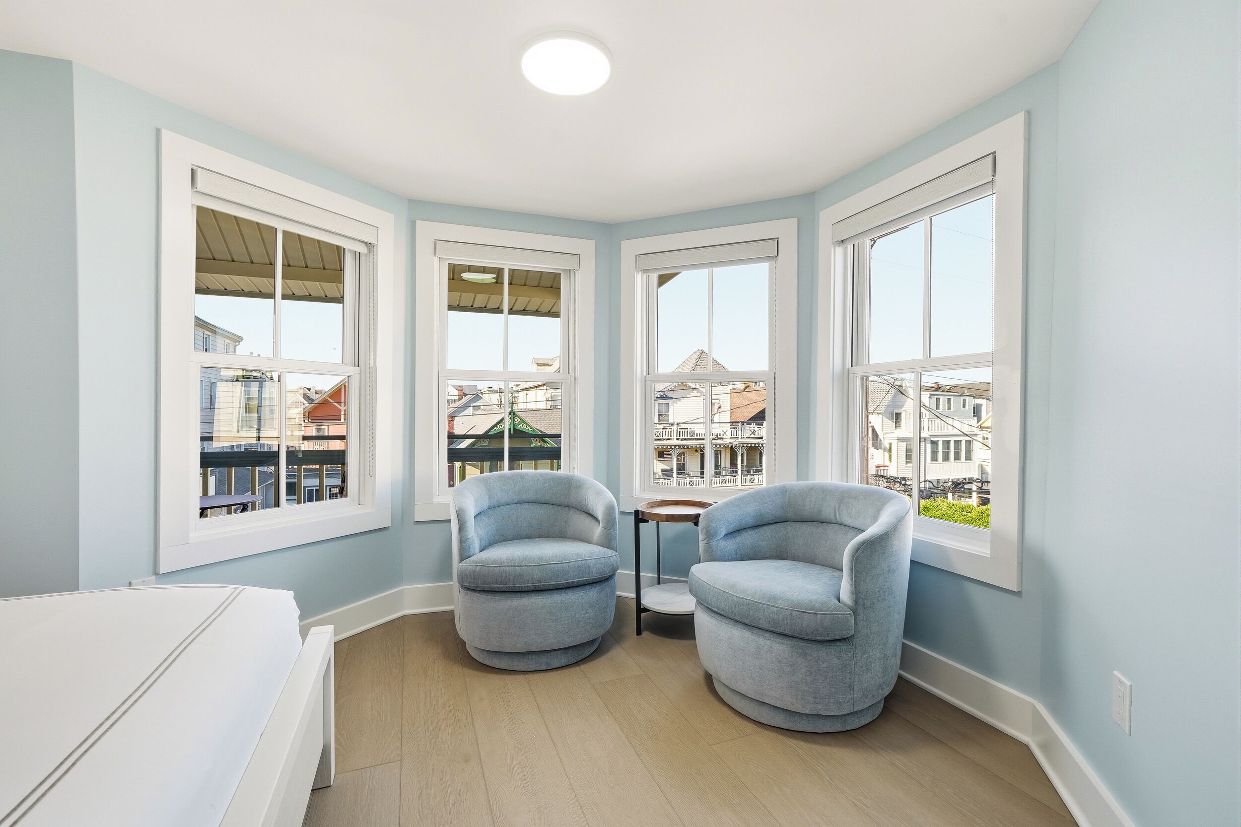 Una habitación de esquina con ventanas amplias, una cama blanca, dos sillones azules y una pequeña mesa.