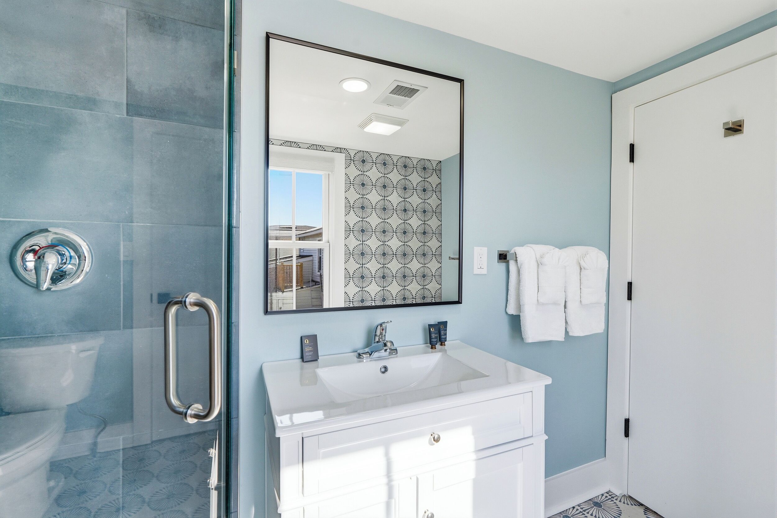 A bathroom with a glass shower enclosure, a white sink with a mirror above it, and a door with a towel rack.