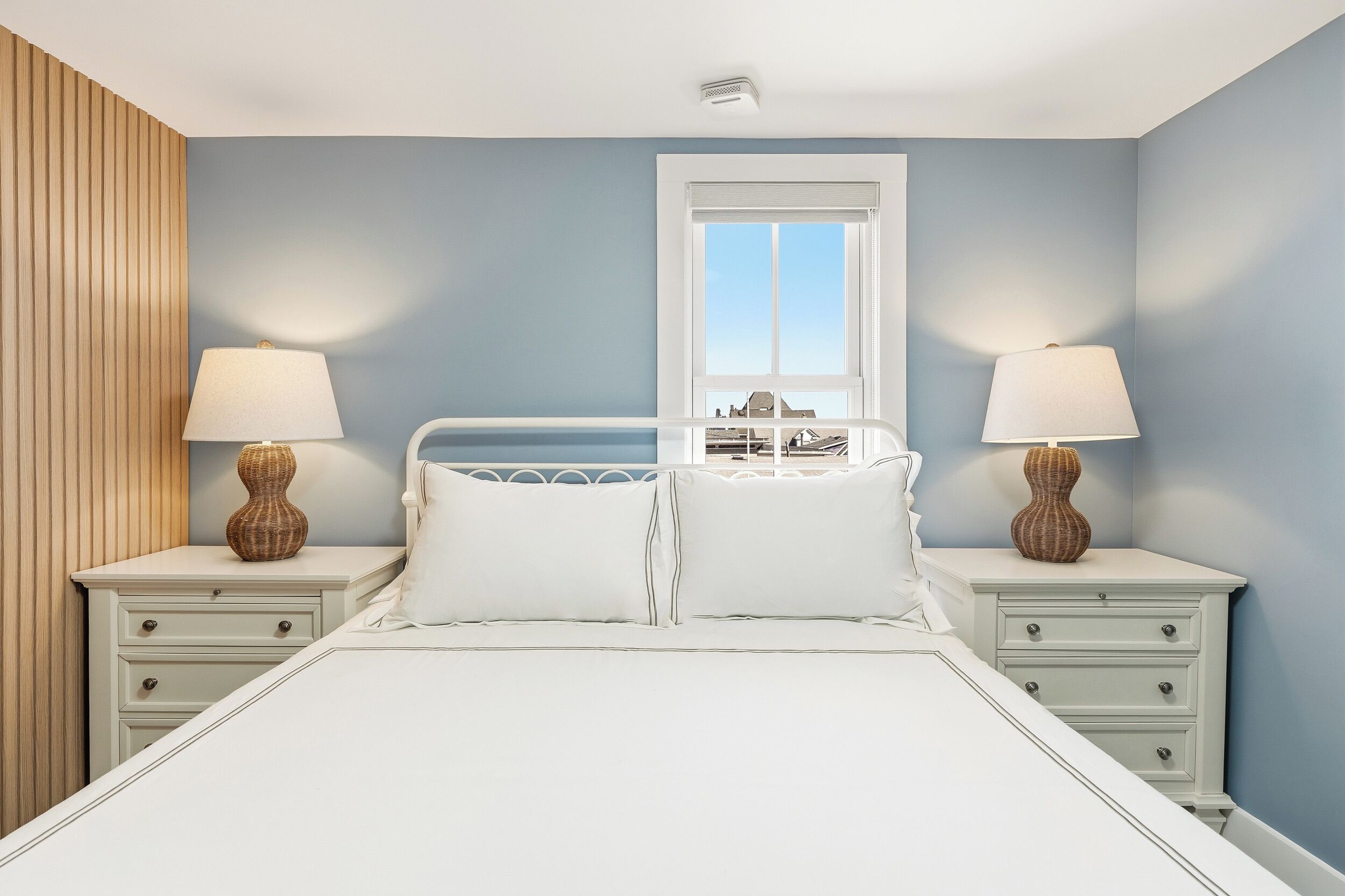 A bedroom with a bed, two bedside tables, a window, and a view of buildings.