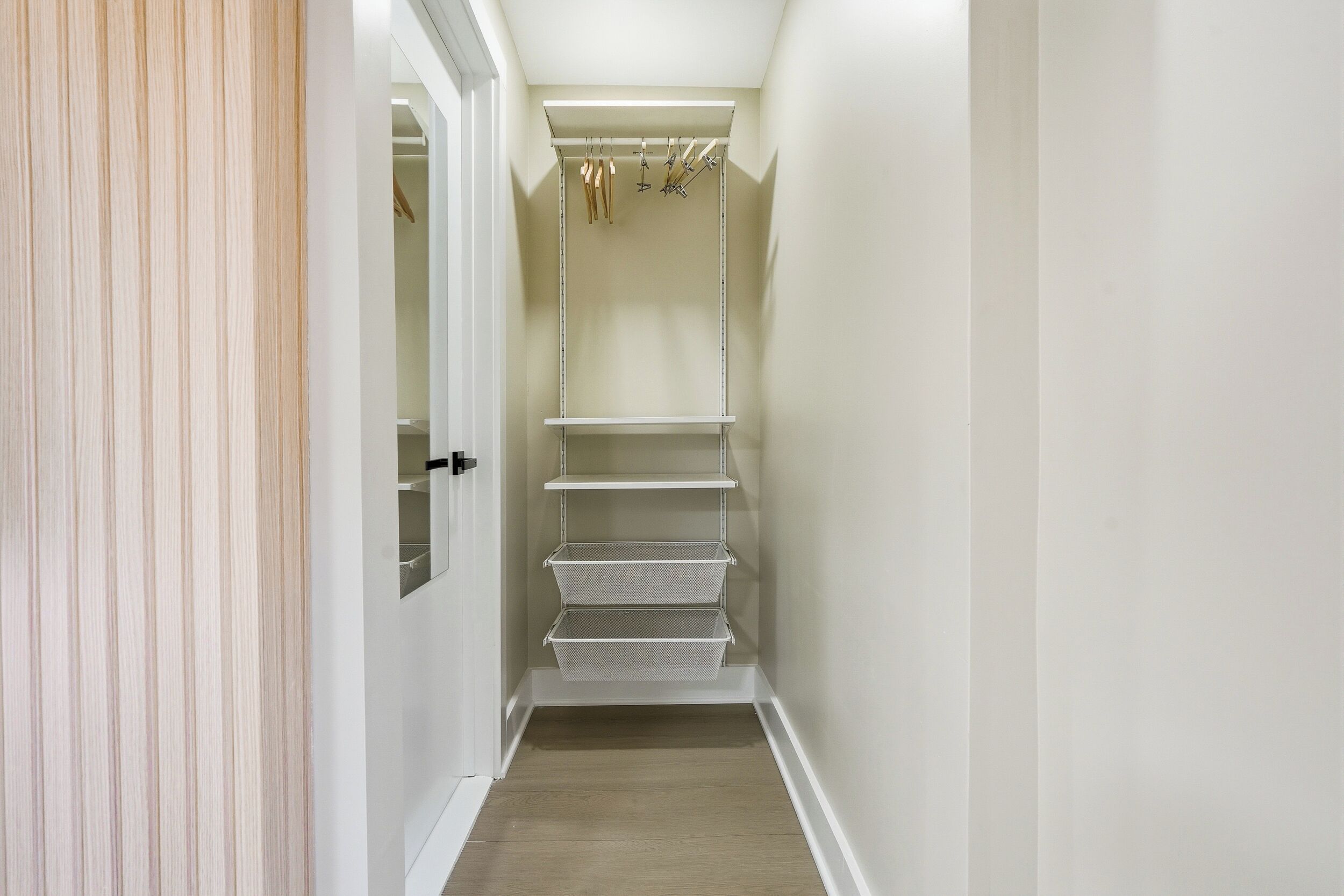 A narrow hallway with a white door on the left, a white closet with shelves and hanging rods, and a wooden floor.