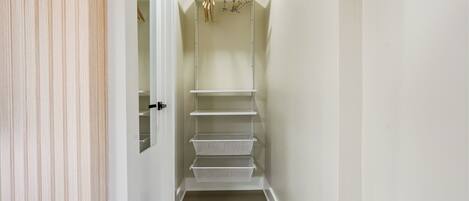 A narrow hallway with a white door on the left, a white closet with shelves and hanging rods, and a wooden floor.