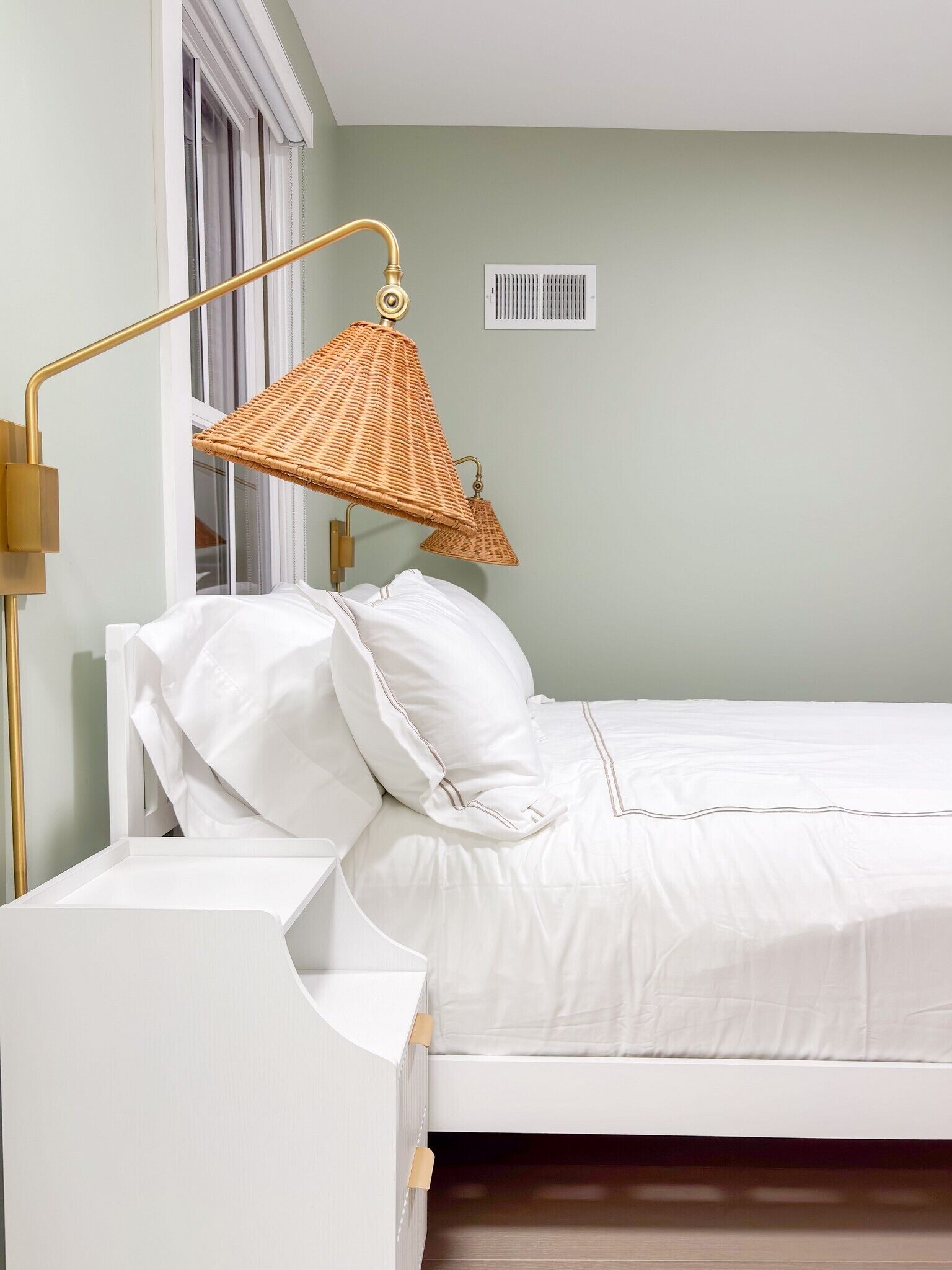 Un lit bien fait, avec une literie blanche, une lampe dorée et un abat-jour en rotin tressé.