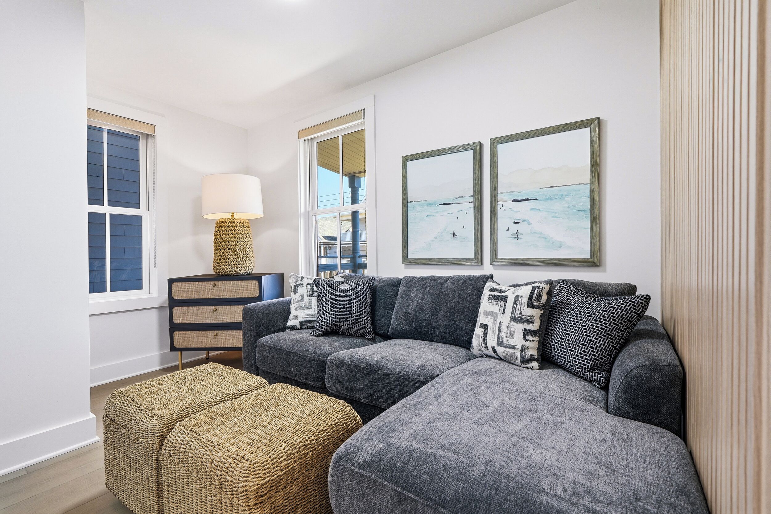 A living room with a gray sectional sofa, two woven ottomans, a lamp, a dresser, and two framed landscape paintings.