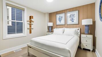 Une chambre à coucher avec un grand lit, deux tables de chevet, une fenêtre avec des stores et des œuvres d’art accrochées au mur.