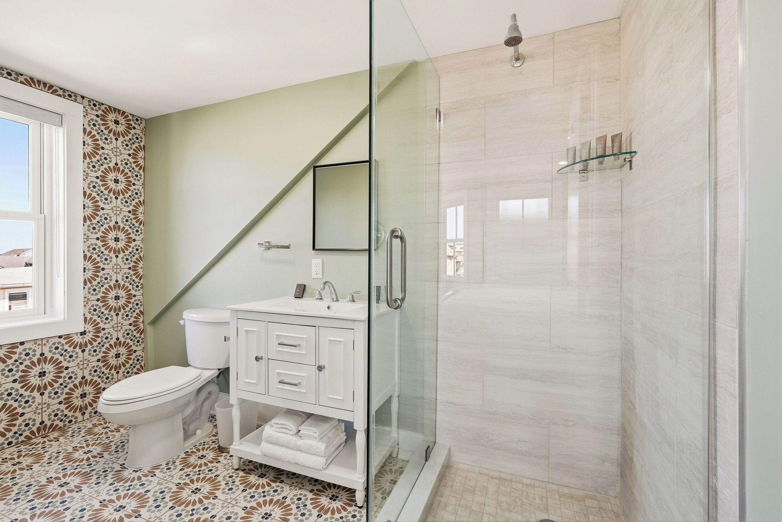 A bathroom with a toilet, white vanity, glass shower enclosure, patterned floor tiles, and a window.