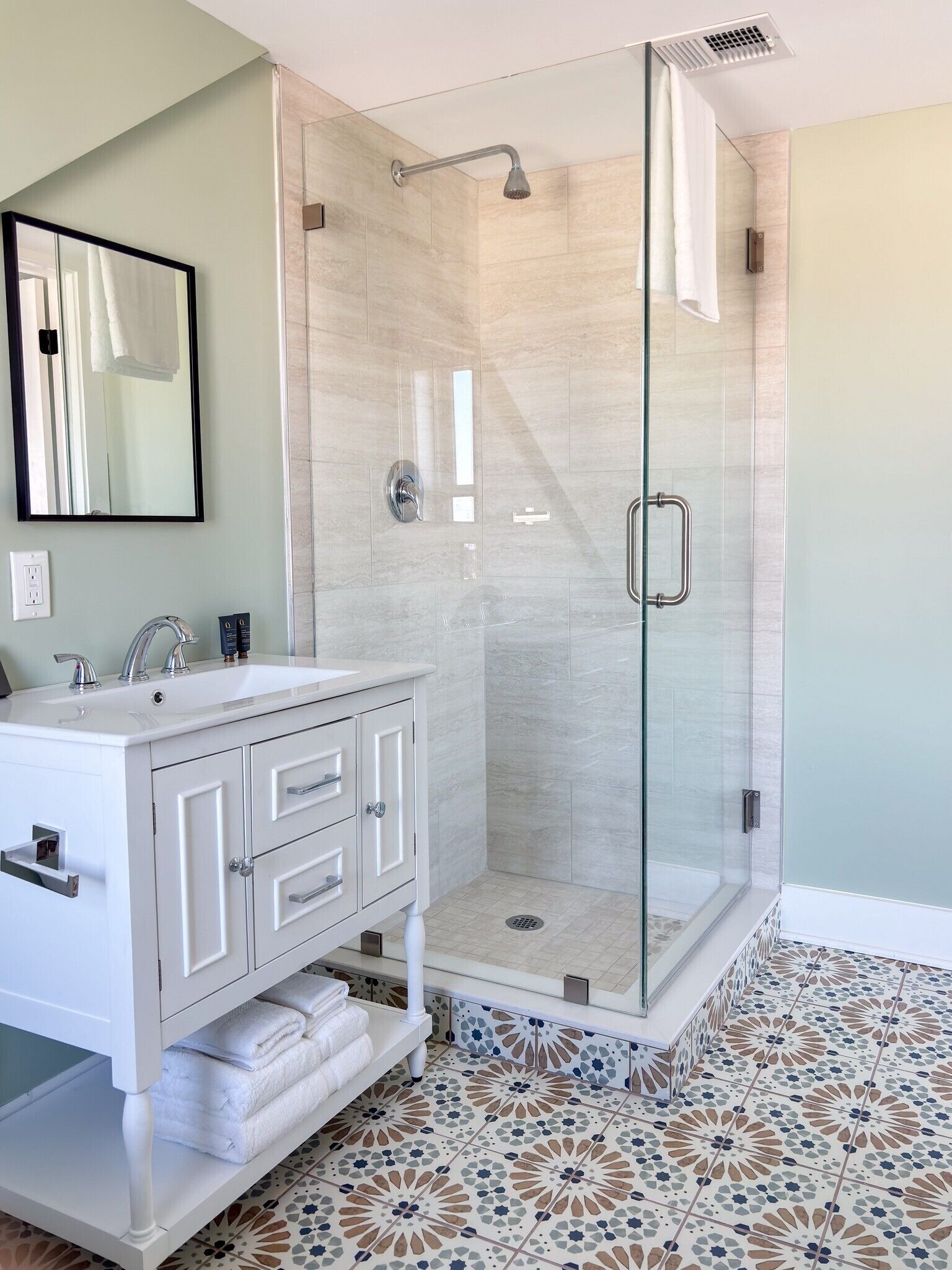 A bathroom with a glass-enclosed shower, a white vanity with drawers, and a patterned floor.