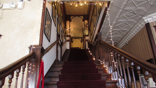 A grand staircase with wooden railings and a red flag, leading to a room with a patterned ceiling and string lights. - The Historic Argo Hotel (Crofton)