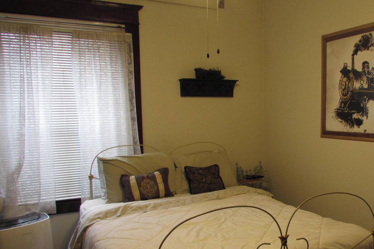 A bedroom with a bed, a window with blinds, a wall-mounted shelf, and a framed picture of a train.