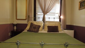 A bed with a metal headboard, two pillows, a bedside table with a lamp, and a window with curtains. - The Historic Argo Hotel (Crofton)